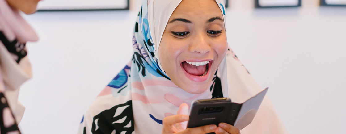 Photo of young woman in headscarf, smiling broadly while looking at smart phone